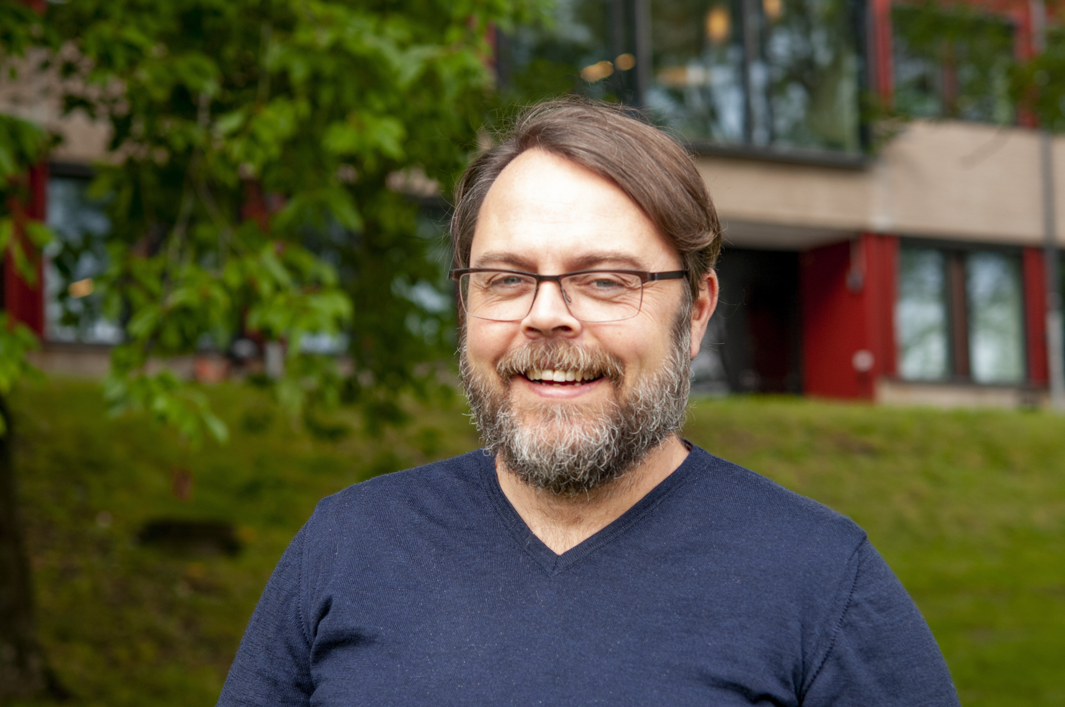 Porträttfoto av en person utomhus. Personen bär glasögon och en mörkblå T-shirt med V-ringning.