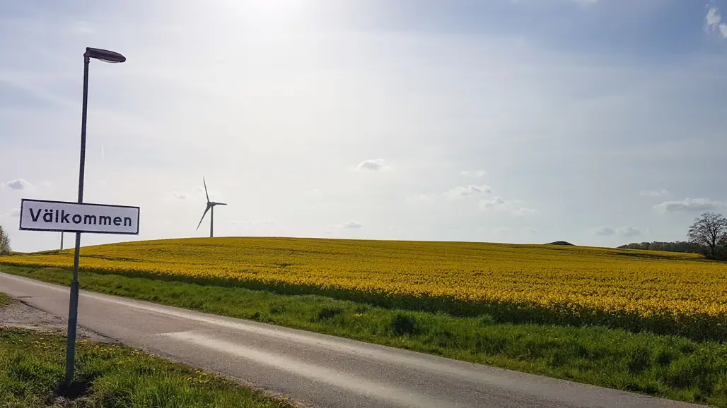 En välkommen-skylt framför rapsfält och vindkraftverk