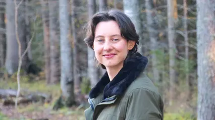 A person with short dark hair and a green jacket stands smiling in a forest with tall trees in the background.