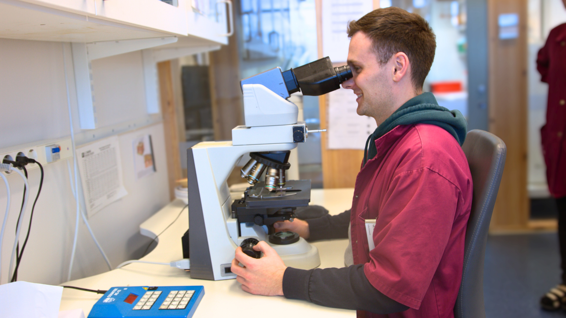Man med vinröd jacka och grön huva som sitter ned framför ett mikroskop. 