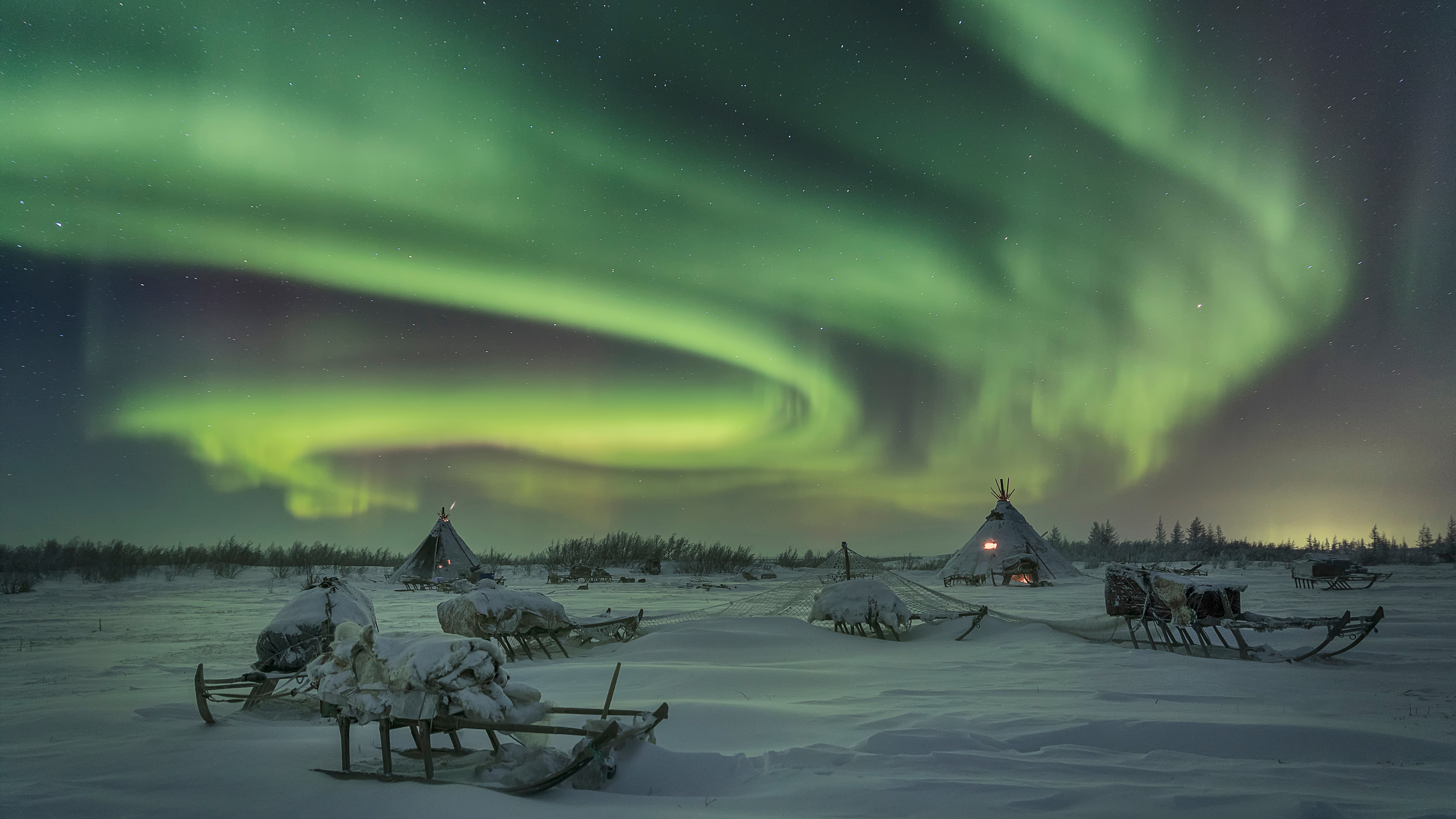Två kåtor och flera slädar i snön. Grönt norrsken på himlen. 