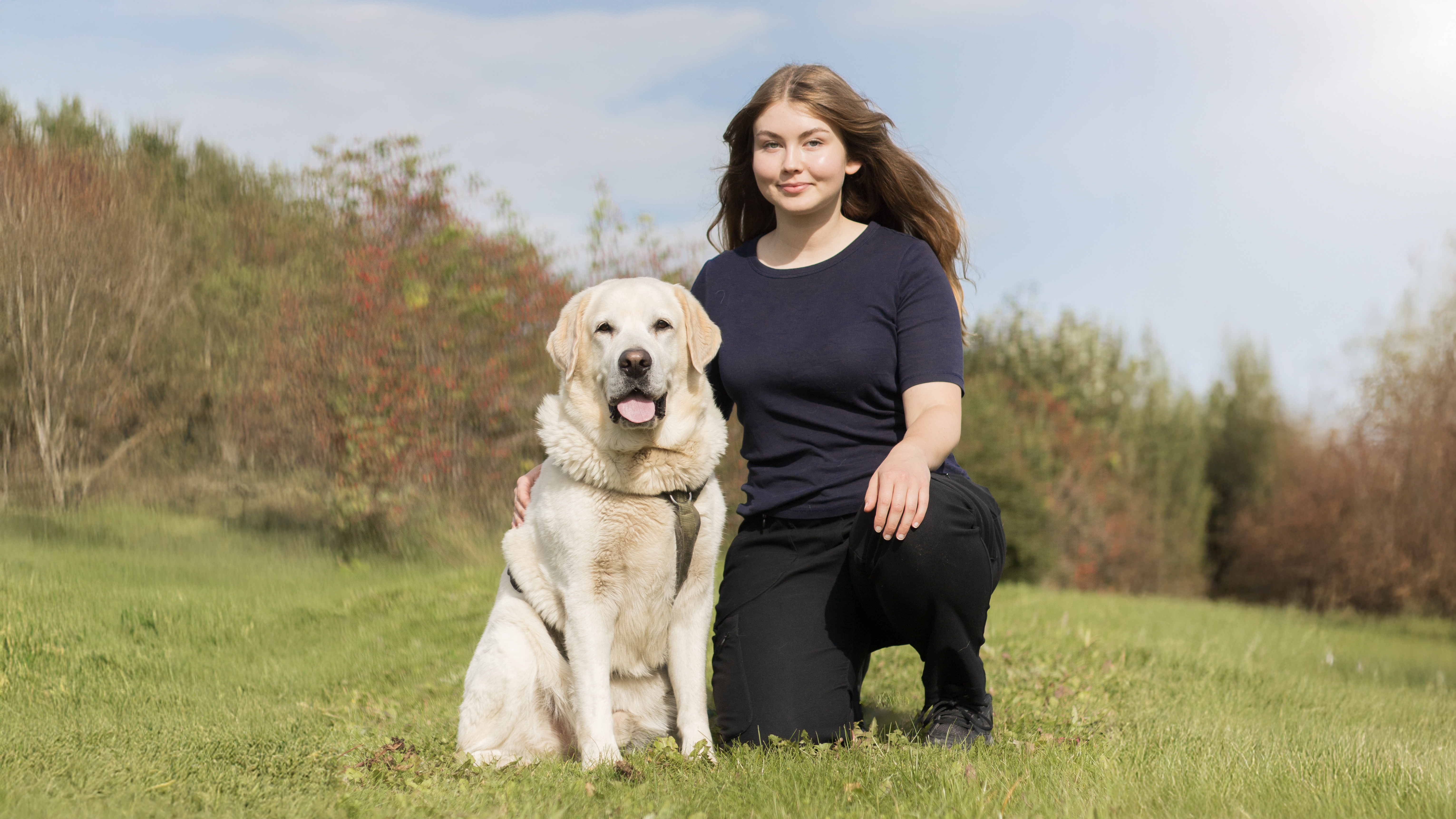 Foto av en kvinna (Felicia Palm, SLU) sittandes på huk utomhus med en ljus labrador retriever.