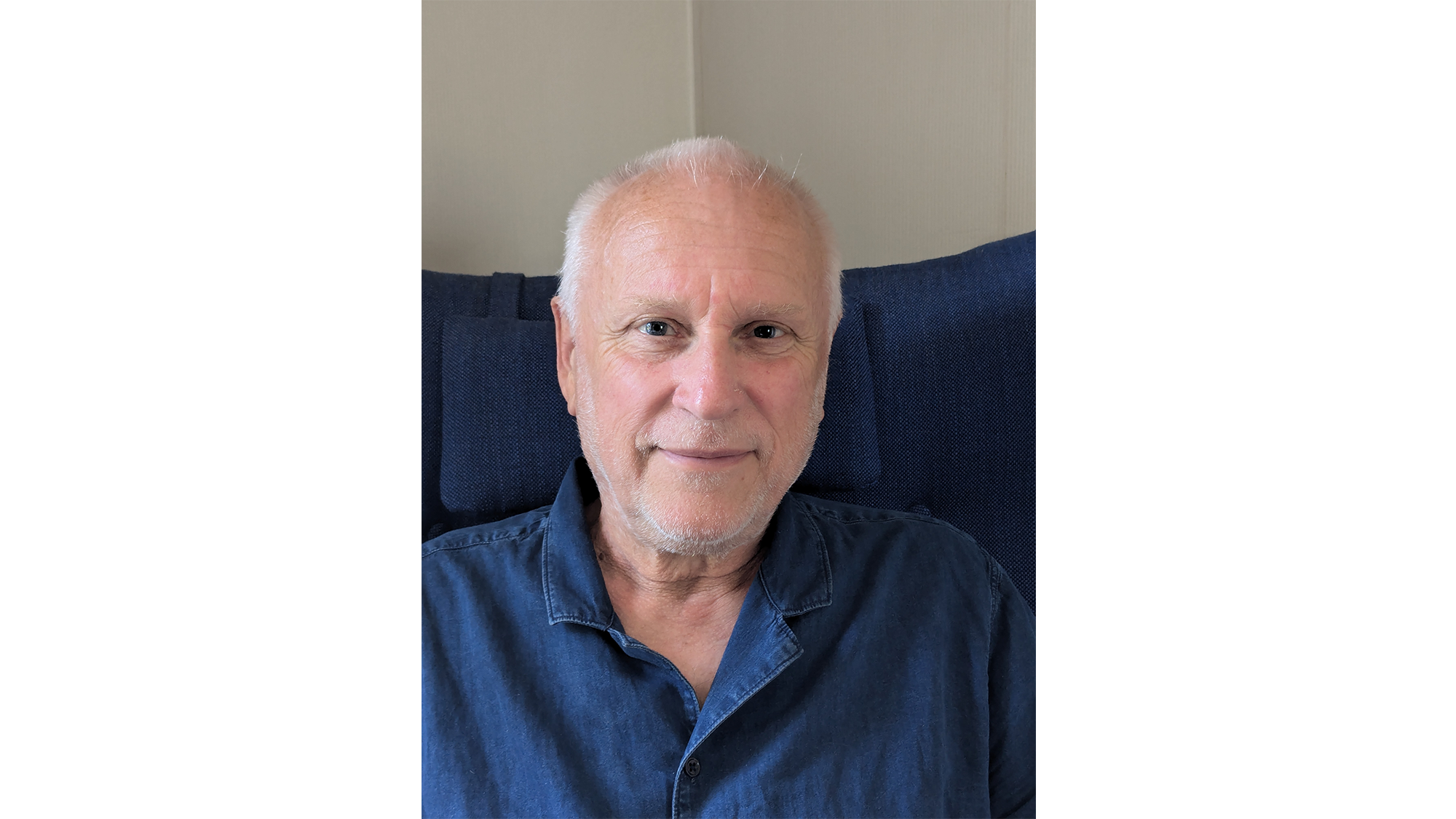 Man med vitt kort hår, vit skäggstubb och mörkblå skjorta sitter i mörkblå fåtölj. Foto