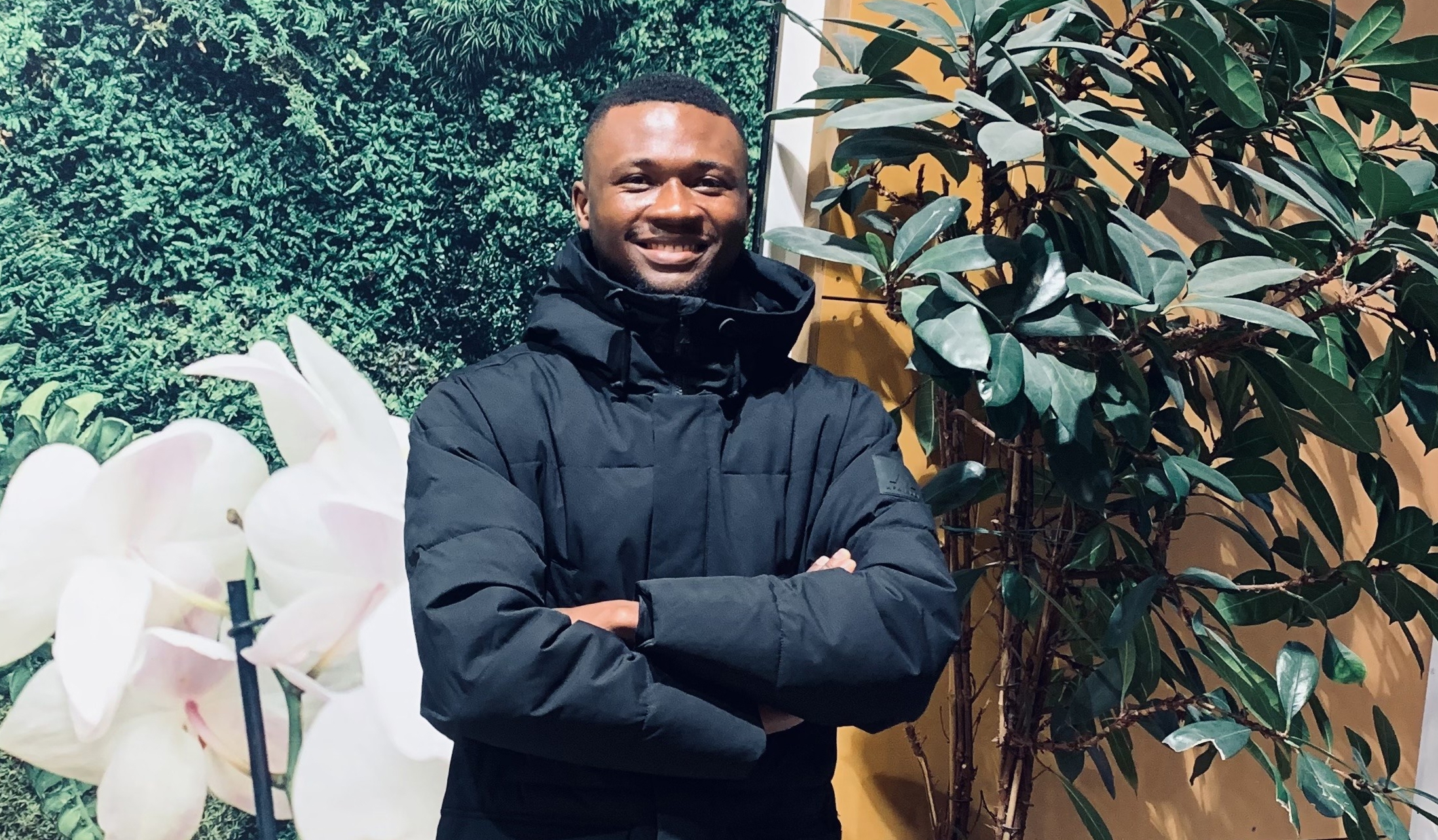 Wisdom, an SLU student ambassador, smiles at the camera in front of a green plant wall.