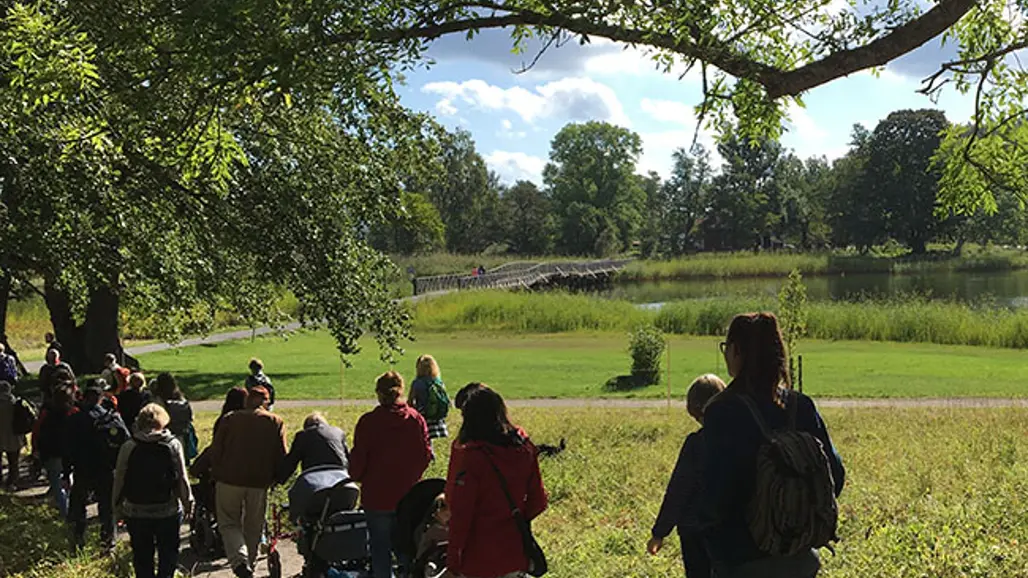 En grupp människor går i Tyresö slottspark en härlig vårdag. Vattnet glittrar i bakgrunden.