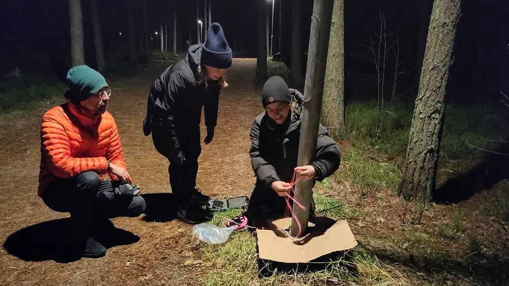 Tre personer på en skogsväg i mörker upplyst av gatulampor. En av personerna mäter ljusstyrkan med ett mätinstument.