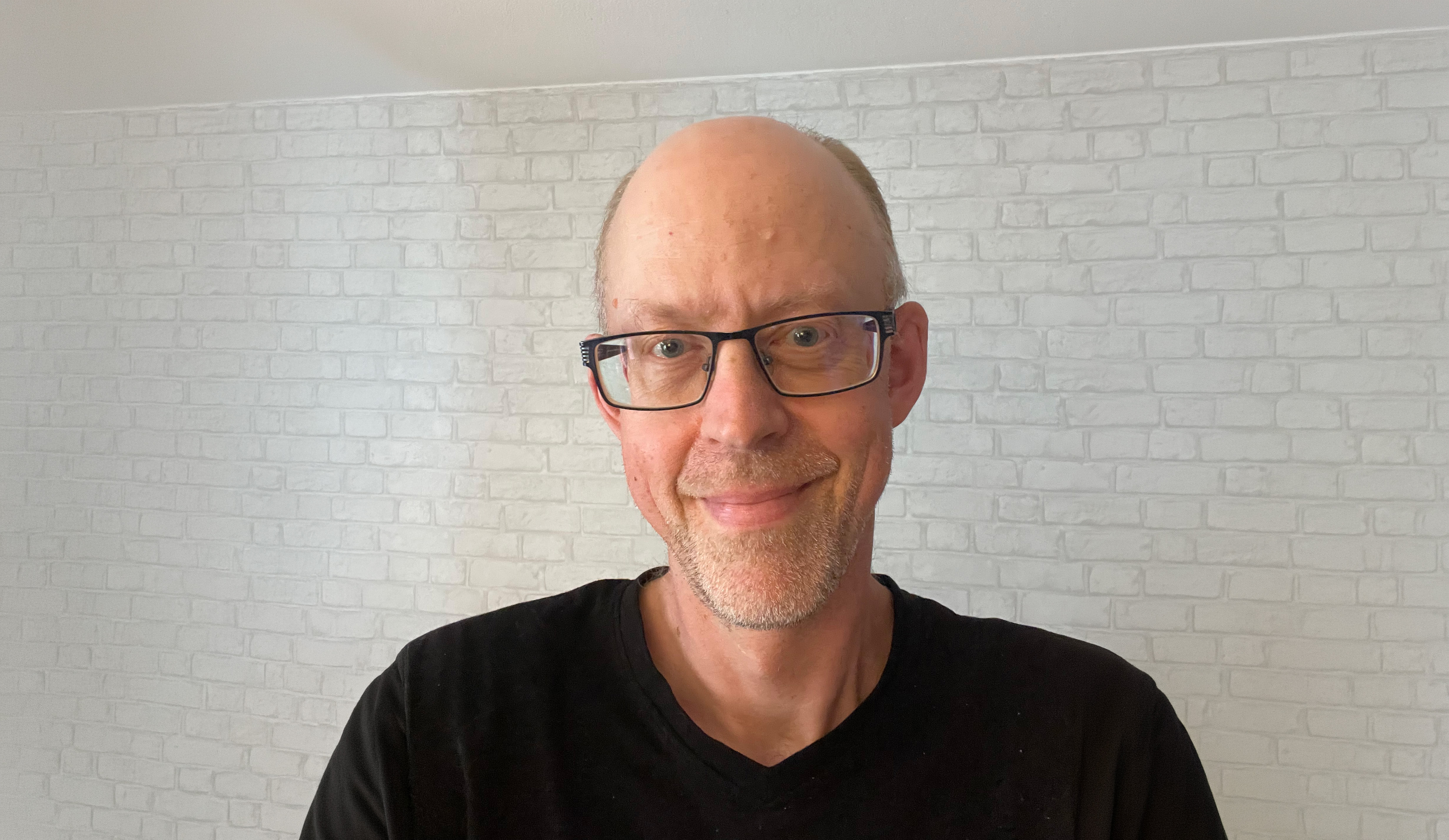Porträttfoto av en person inomhus framför en ljus vägg med tegelmönster. Personen bär glasögon och en svart T-shirt.