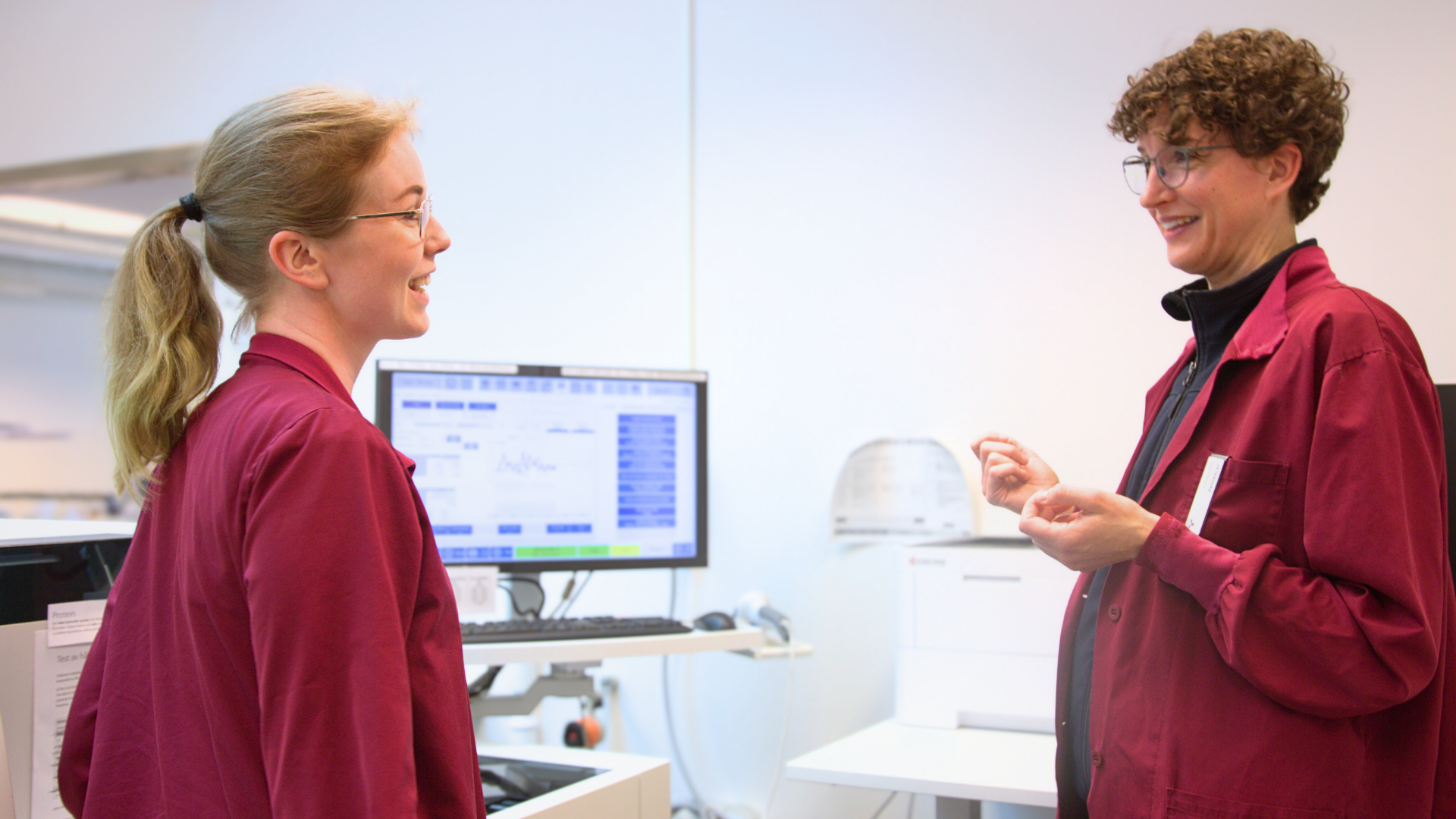 Two women in burgundy clothes standing next to each other. A computer screen is visible in the background.