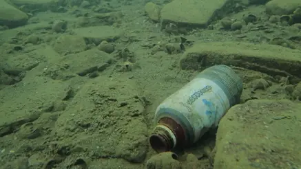 Underwater scene showing a discarded plastic bottle on a rocky seabed, surrounded by scattered shells.