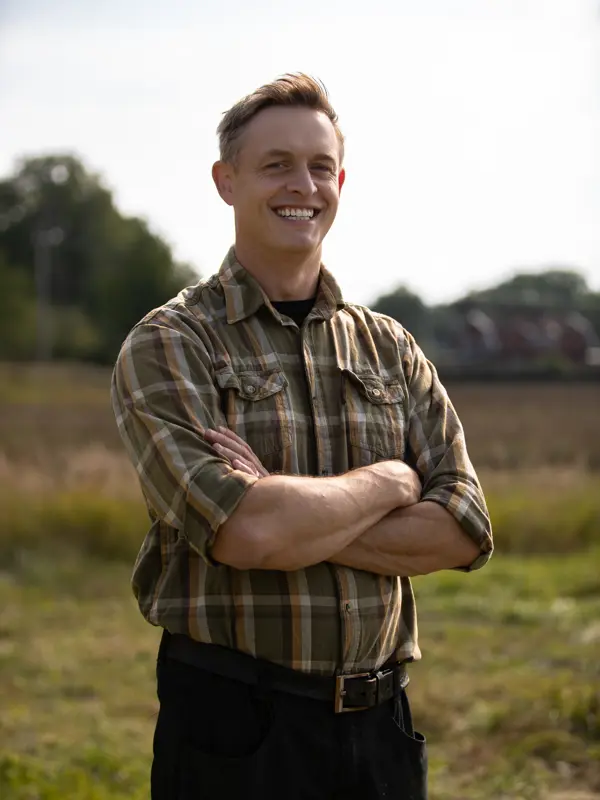 Leende man i rutig skjorta står med armarna i kors på en åker i kvällsljus.