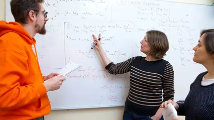 Tre forskarstuderande diskuterar matematiska formler vid en whiteboard fylld med ekvationer. En person pekar på tavlan med en penna medan de andra två lyssnar och håller i anteckningar.