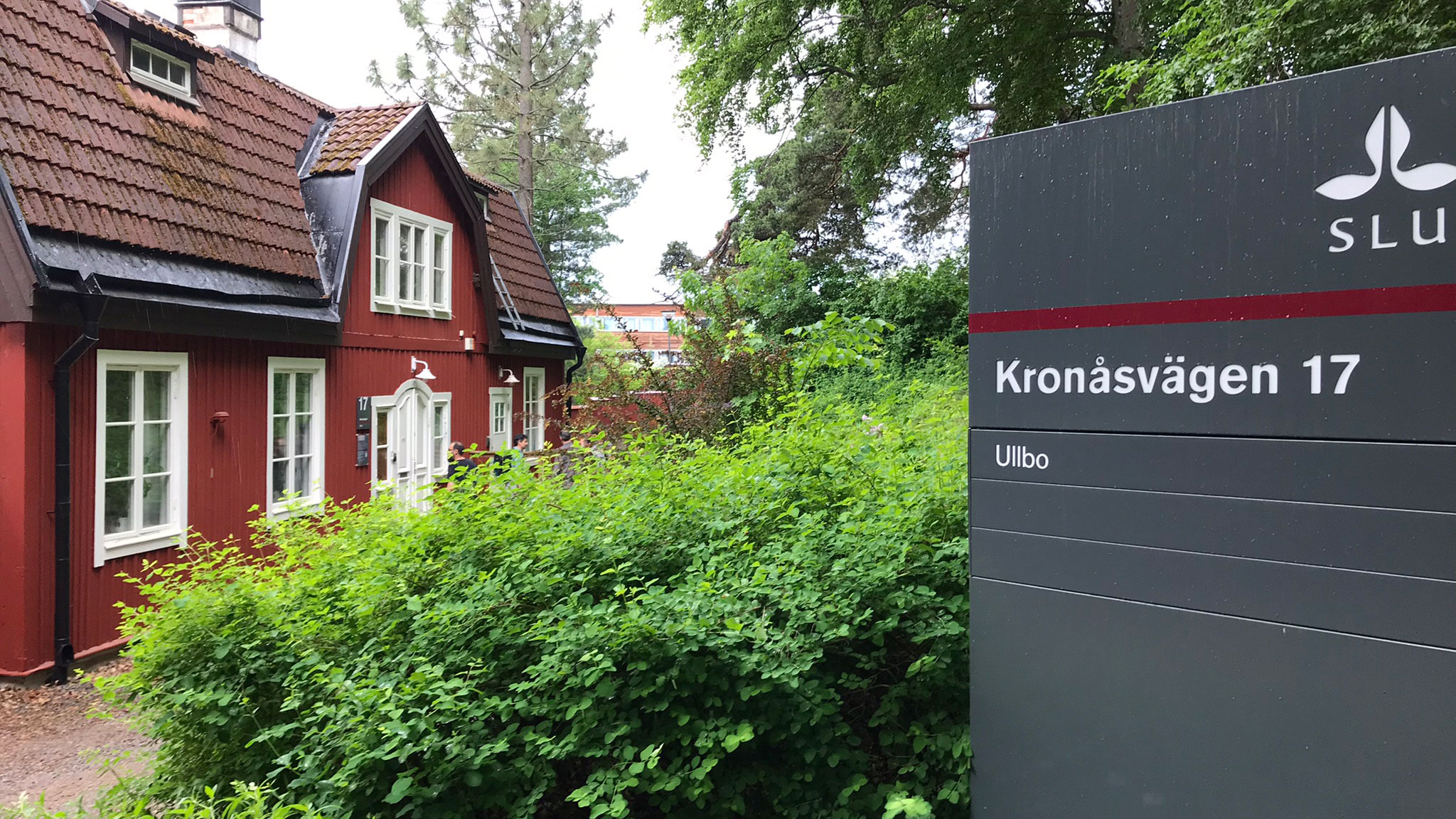 Rött hus med vita knutar, gröna buskar och en skylt utanför. Foto.