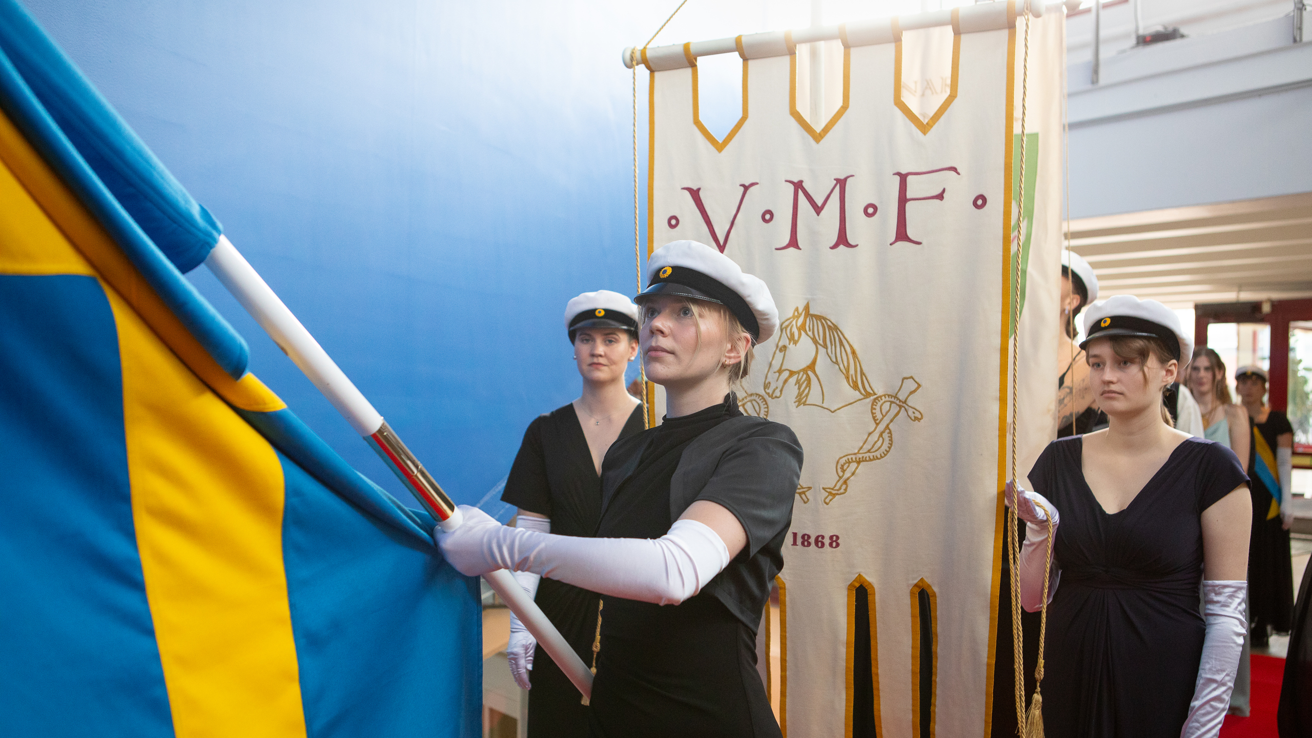 Studenter i svarta högtidskläder, vita handskar och studentmössor bär fanor i procession.