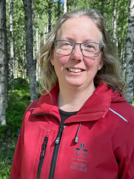 A portrait of a person wearing a red jacket and glasses, standing in a forest.