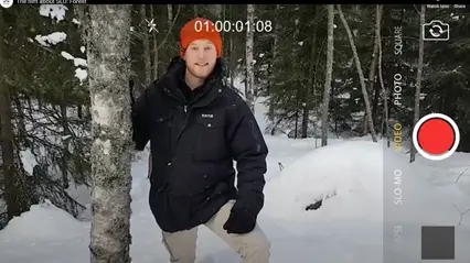 En person i svart jacka och orange mössa står i en snöig skog och lutar sig mot en björk. Bilden är en stillbild ur en video, vilket syns på skärmelement som en röd inspelningsknapp, tidskod och kamerameny till höger i bild. Granar och snötäckt mark omger personen.