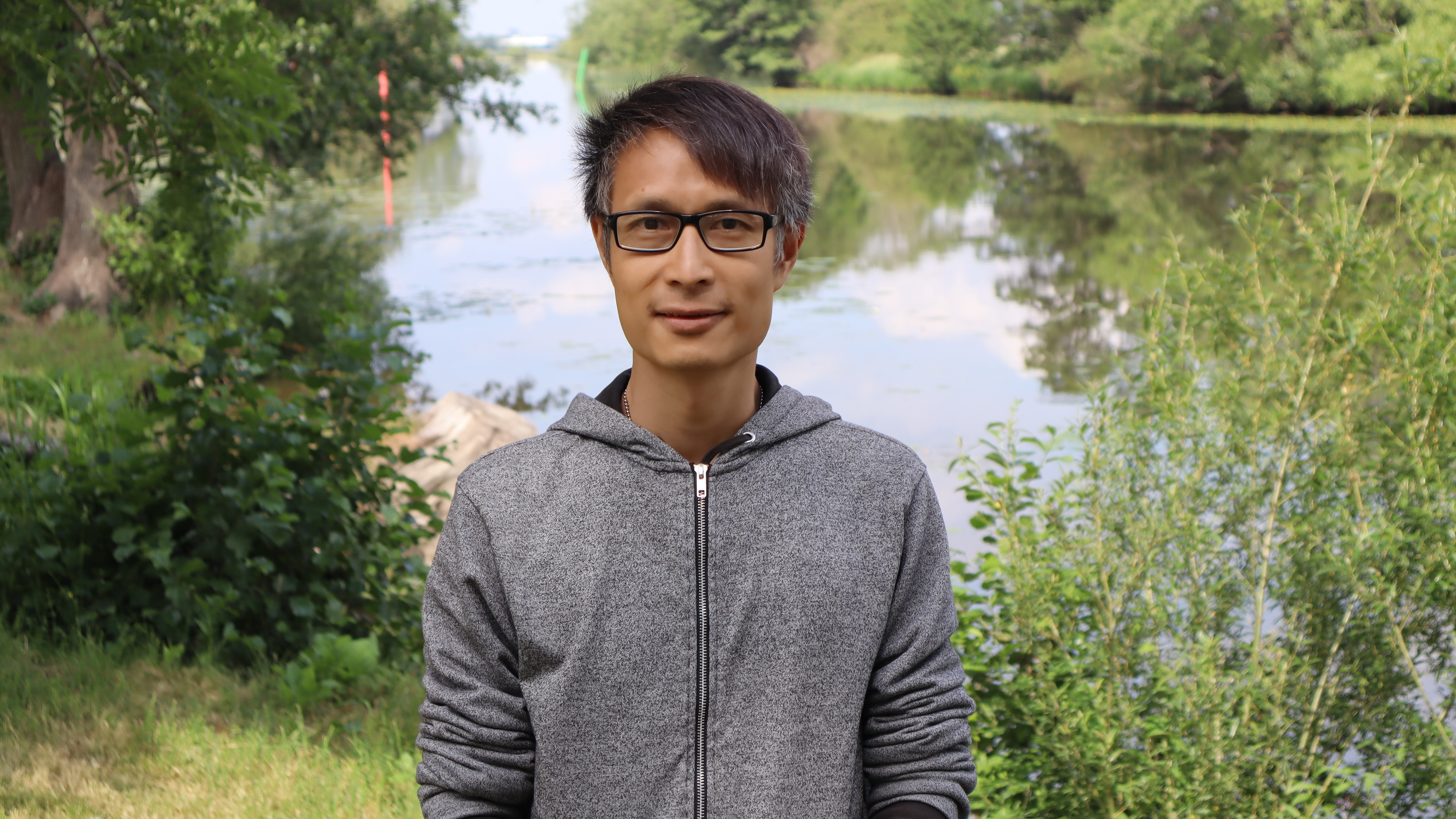 Porträtt av en man med glasögon och grå luvtröja, utomhus framför en sjö och grönska.