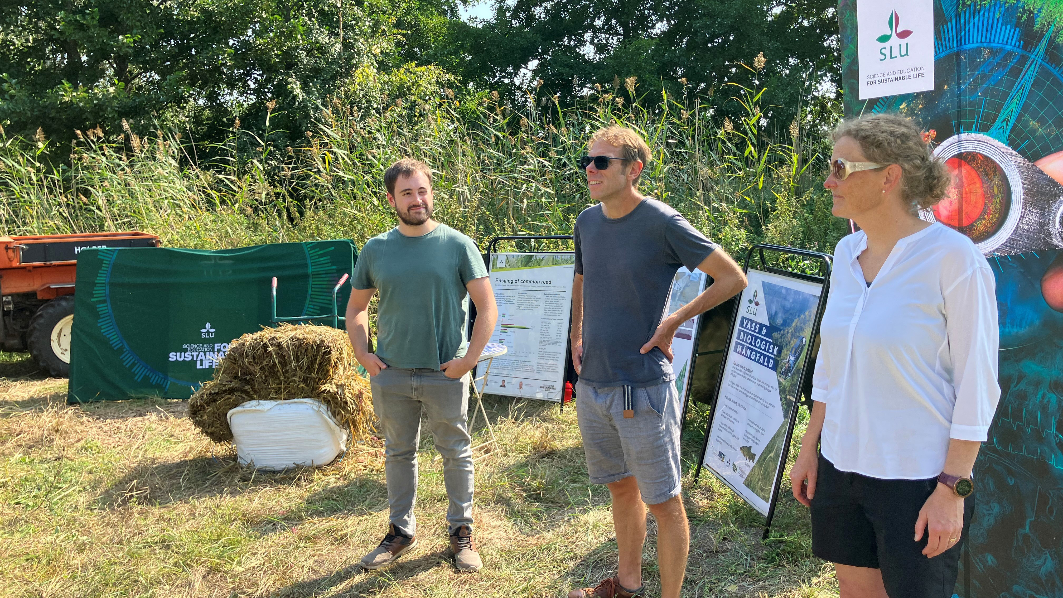 William Ashworth, Örjan Östman och Malin Connysson framför SLU:s monter, utomhus med vass och träd i bakgrunden. Foto. 