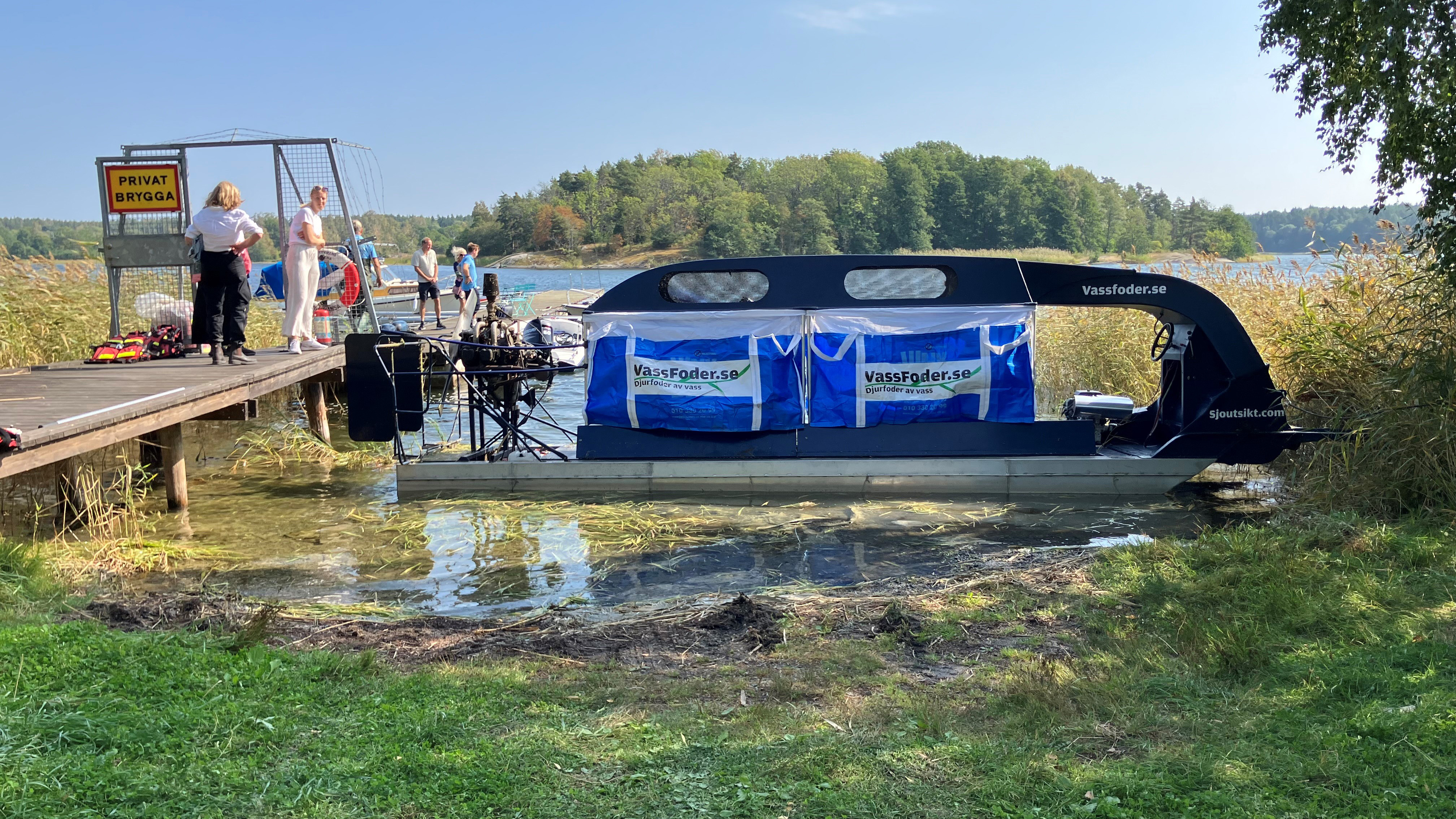 Vasskördaren ligger förtöjd i en vik bredvid en brygga med några människor på. Foto. 