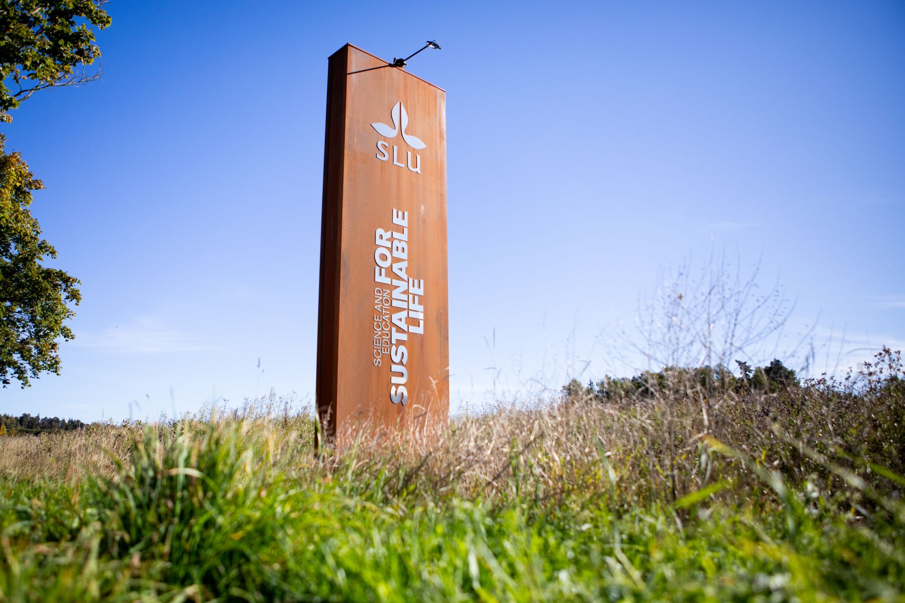 SLU sign reading “Science and Education for Sustainable Life” in an open landscape.