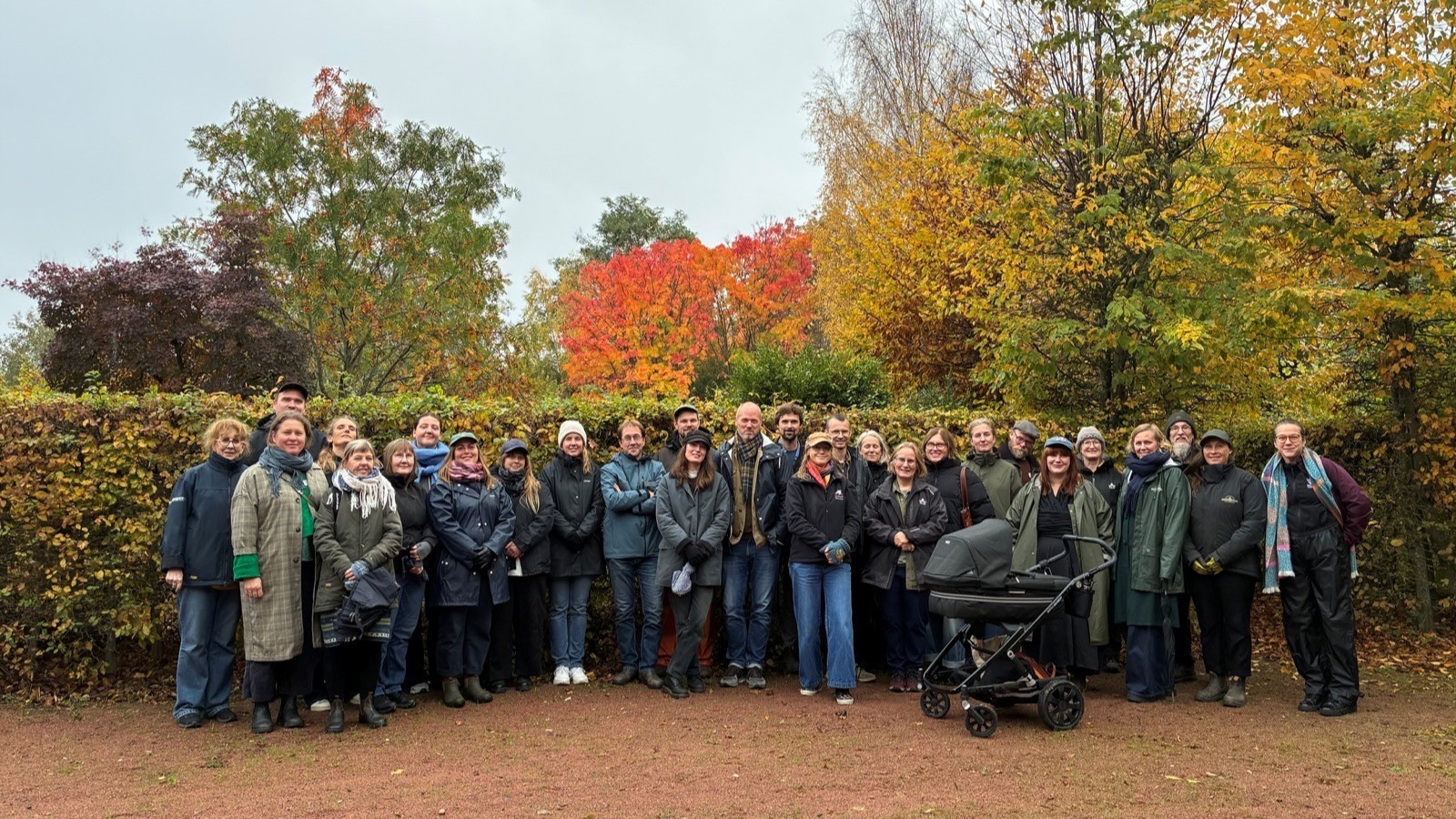 Ett gruppfoto av genbankens personal och de klonarkivsvärdar som deltog på träffen. Ett trettiotal personer, många klädda i grönt, svart och marinblått, står framför en bokhäck. Det är mulet och regnet hänger i luften. 