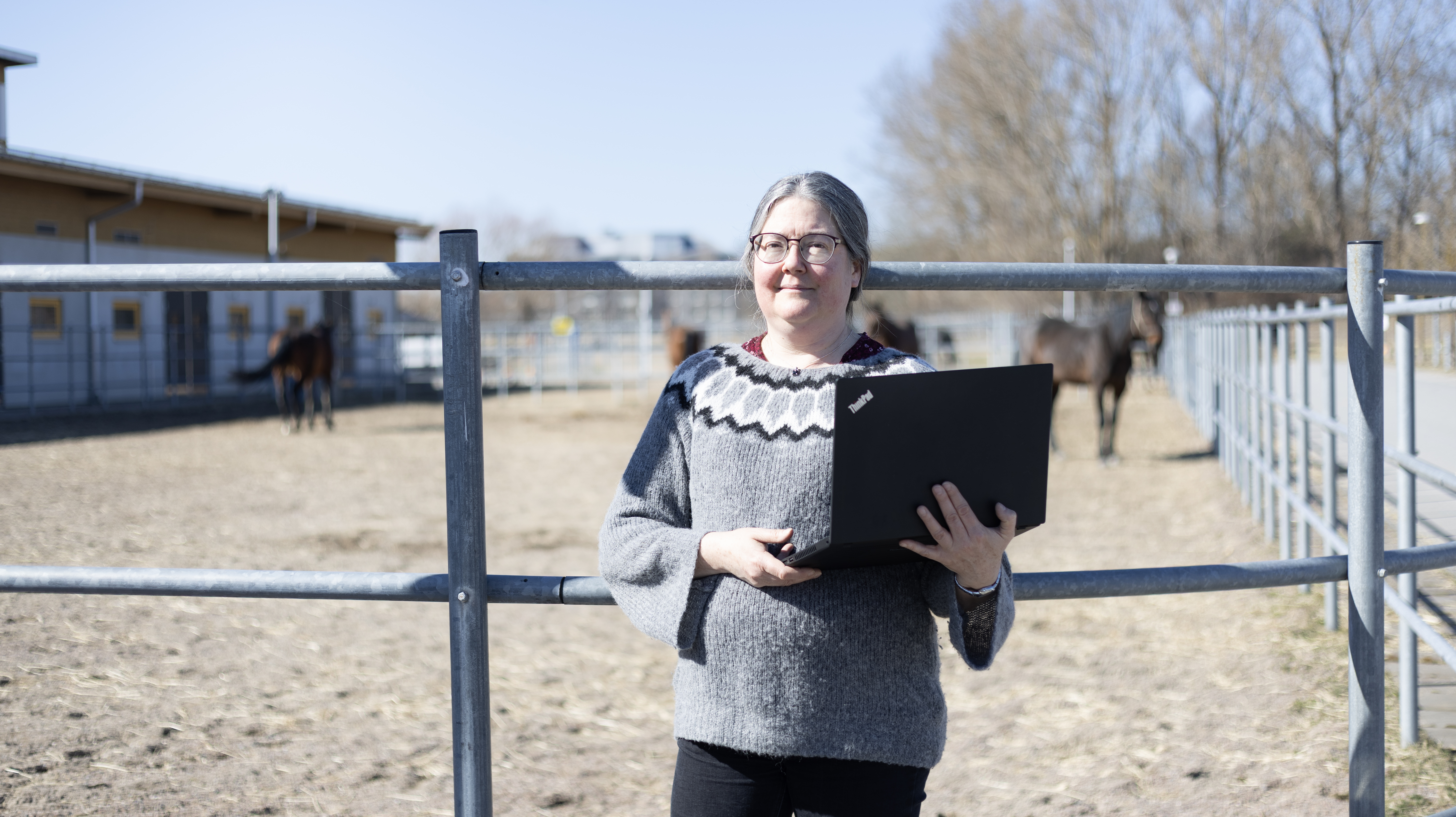 Photo of a woman (Professor Susanne Eriksson, SLU) standing with a laptop in her arms in front of a paddock with horses.
