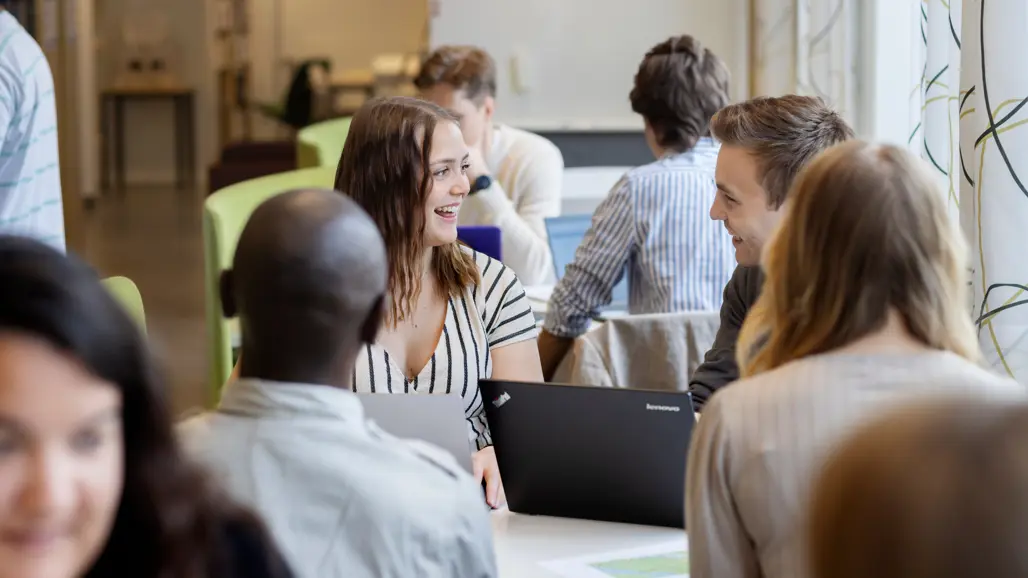 En grupp studenter sitter vid ett bord i ett ljust rum med stora fönster. De arbetar vid sina bärbara datorer och skrattar tillsammans. Fokus är på två personer som ler och samtalar, omgivna av andra i bakgrunden.