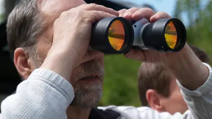 An older man with grey hair and a beard is looking through binoculars with orange-tinted lenses. He is outdoors in daylight with green trees in the background, and another person is partially visible behind him.