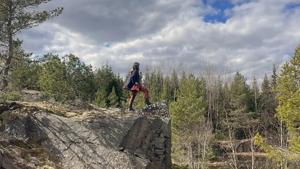 En person står på en klippavsats i naturen, omgiven av träd och under en delvis molnig himmel. Personen bär friluftskläder och verkar njuta av utsikten från den höga platsen. Foto.