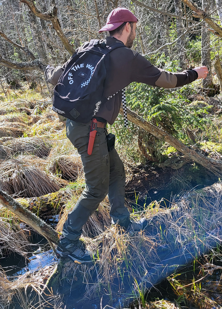 En person med en röd keps och en svart ryggsäck med texten 'OFF THE WALL' går över en smal träbro i en tät skog. Personen bär mörka kläder och har ett rött föremål fäst vid byxorna. Skogsmarken är täckt av gräs och mossa. Foto.