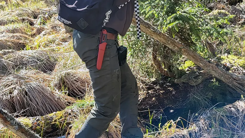 En person med en röd keps och en svart ryggsäck med texten 'OFF THE WALL' går över en smal träbro i en tät skog. Personen bär mörka kläder och har ett rött föremål fäst vid byxorna. Skogsmarken är täckt av gräs och mossa. Foto.