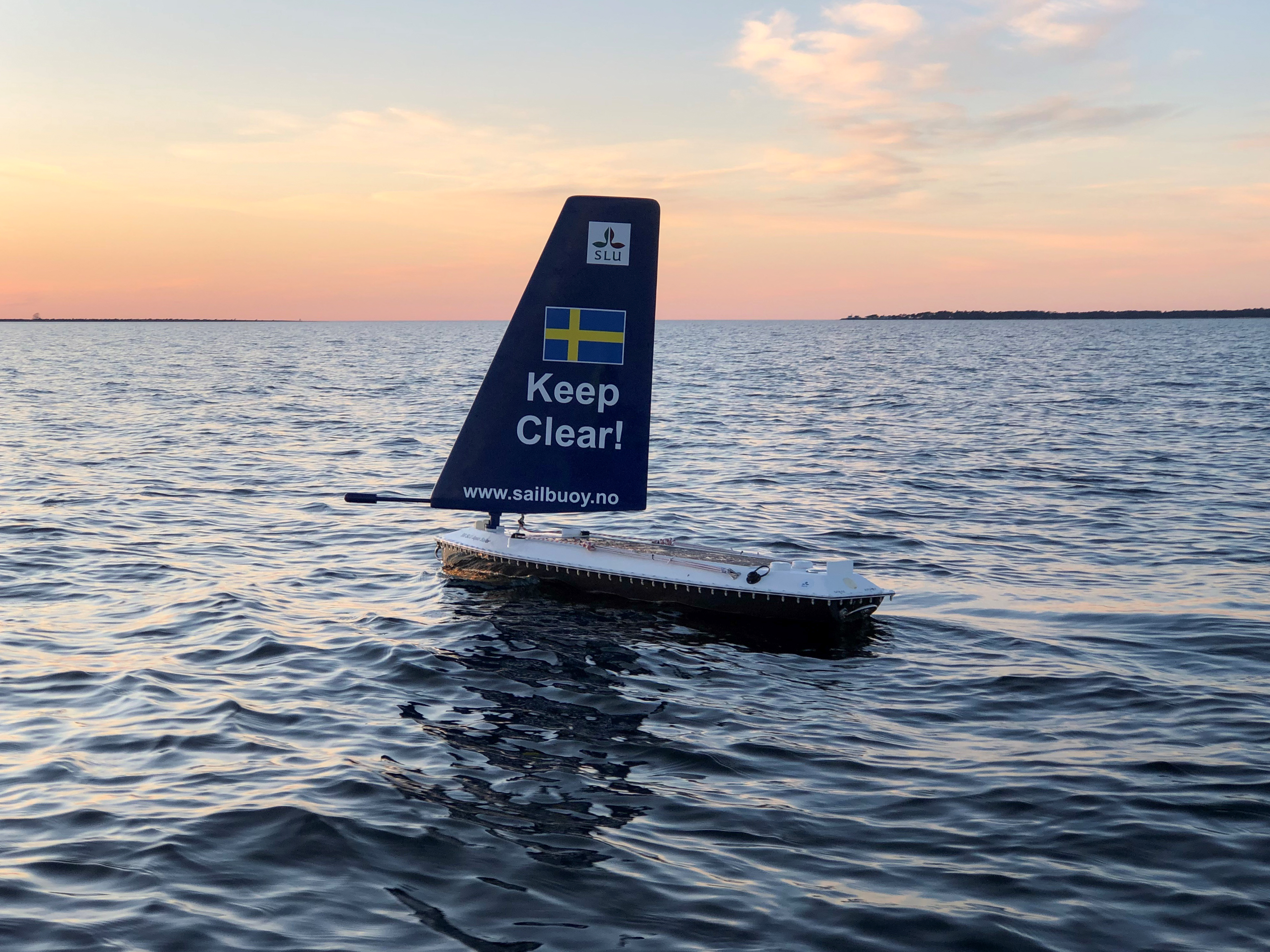 En autonom seglande boj med en blå segelfena som har texten 'Keep Clear!' och en svensk flagga, flyter på en vågig havsyta. Vattenstänk syns i förgrunden, och i bakgrunden syns en kustlinje med byggnader och vindkraftverk under en klar blå himmel.
