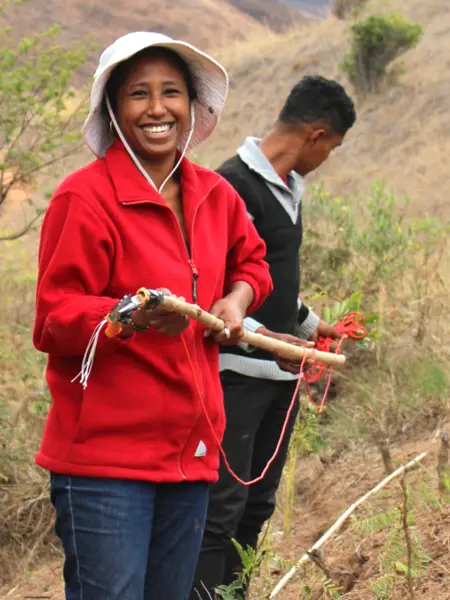 A woman in a red jacket and white hat smiles while holding a tool in her hand and standing on a dry slope. A man works in the background.