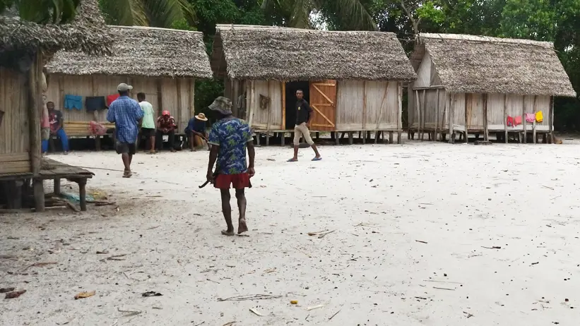 An area with several wooden houses with thatched roofs, and people walking and gathering nearby in a tropical environment.