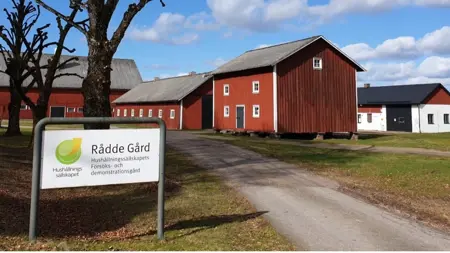 Infarten till Rådde gård, en grusväg med röda lador runt. På en vit skylt står "Rådde gård Hushållningssällskapets försöks- och demonstrationsgård".