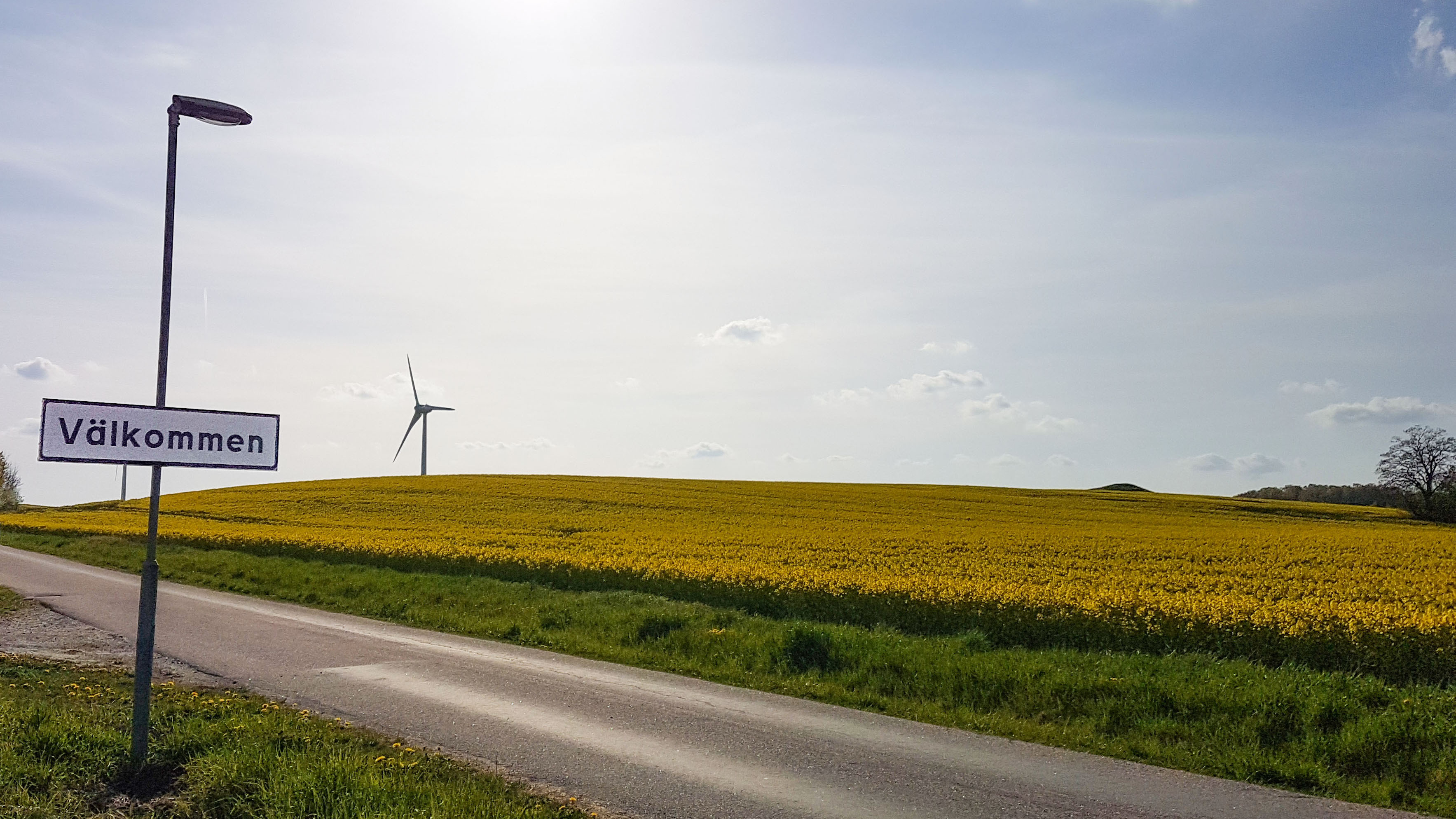 En välkommen-skylt framför rapsfält och vindkraftverk