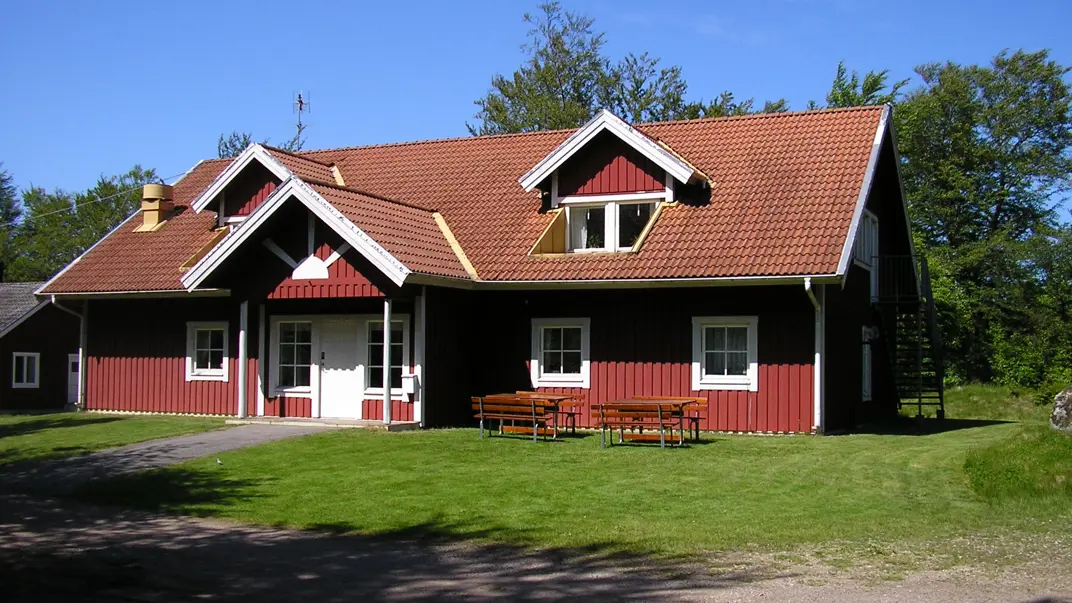 A read two story wooden house with a tile roof and white corners. On the lawn in front of the house are two picknick tables