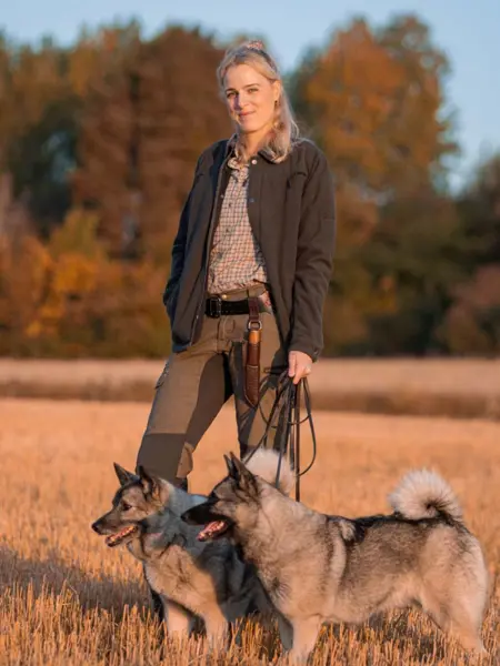 Porträttbild på Josefine Jerlström i jaktkläder ute på en åker med sina två hundar.