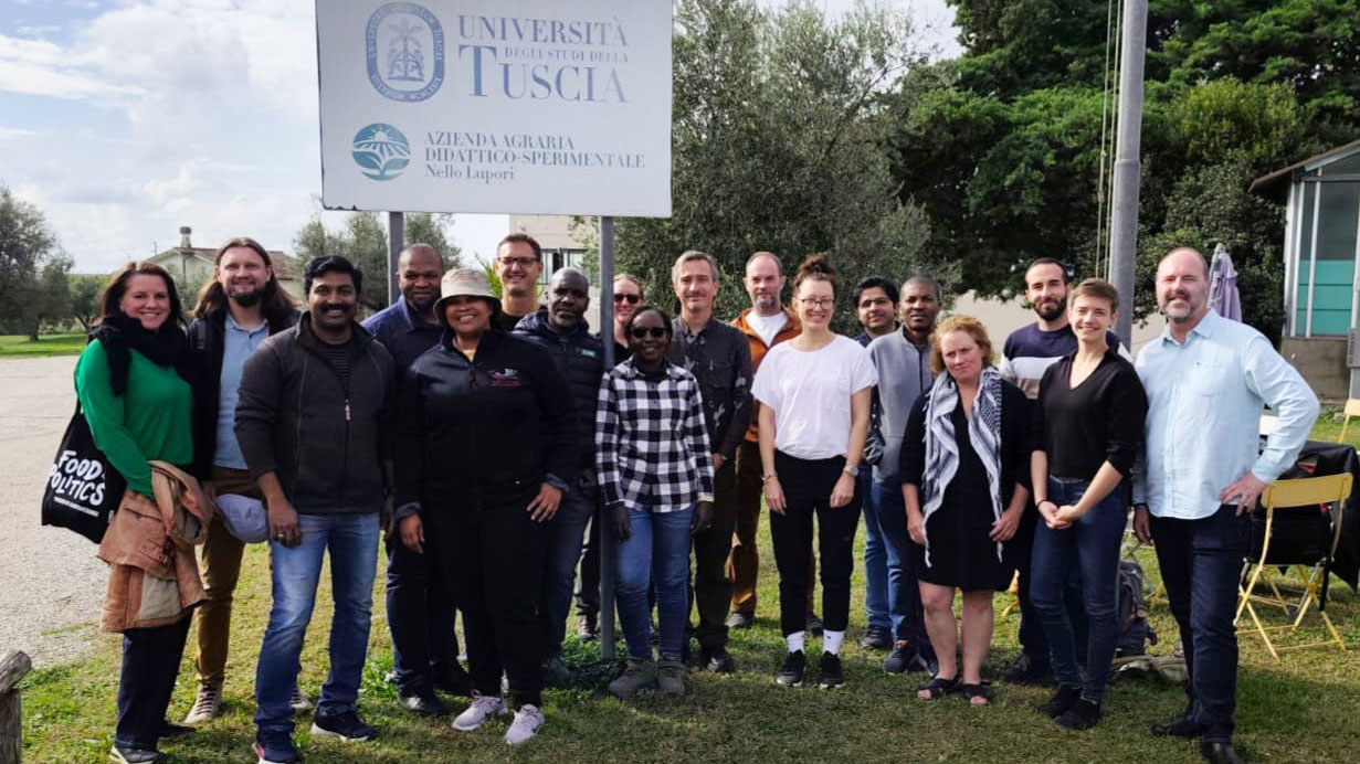 A large group of people standing on a lawn with a sign in the background with the text University of Tuscia. Photo