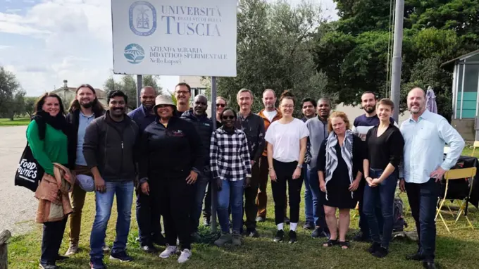 A large group of people standing on a lawn with a sign in the background with the text University of Tuscia. Photo