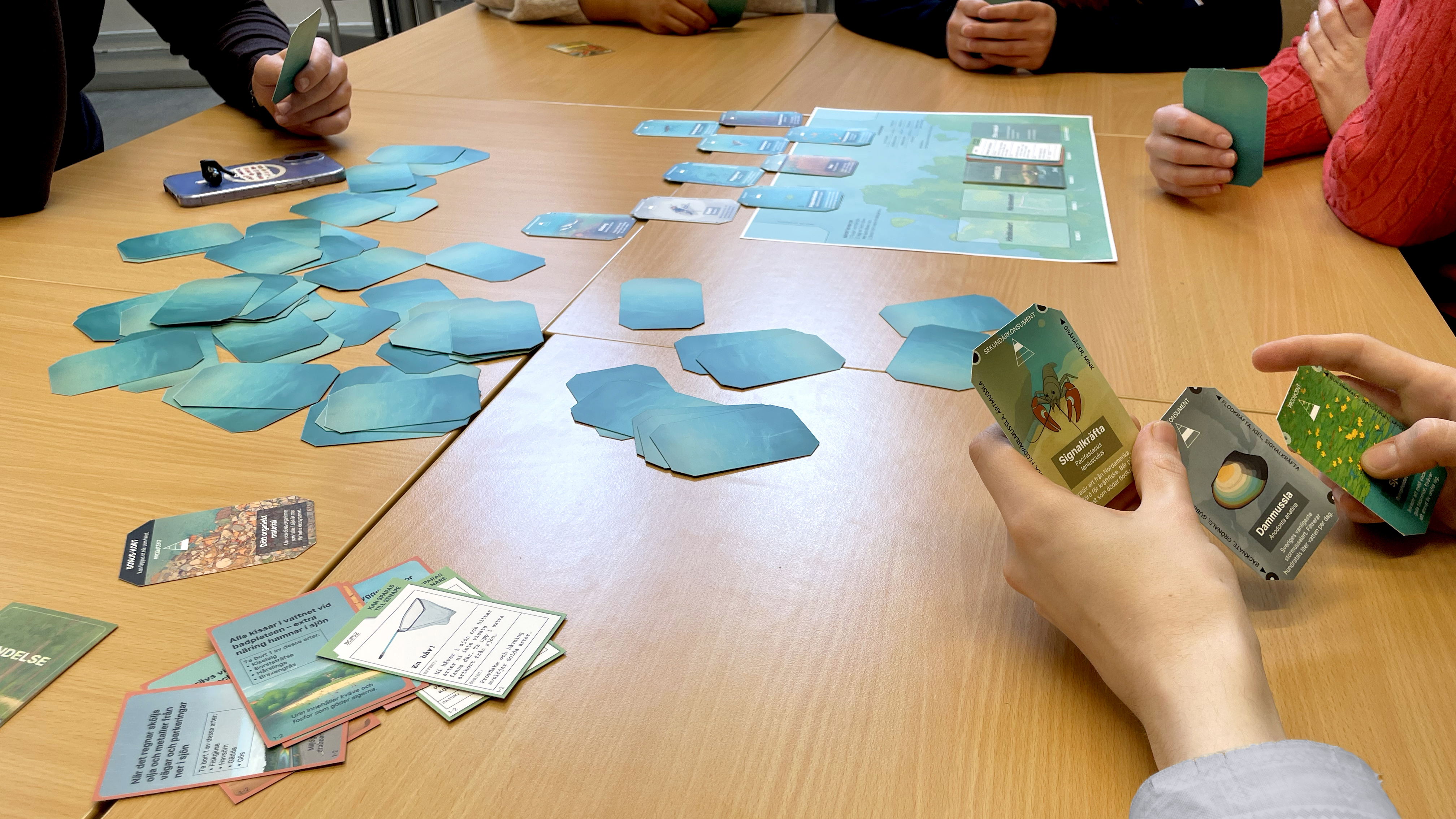 Händer håller spelkort runt ett bord med spelplan. Foto.