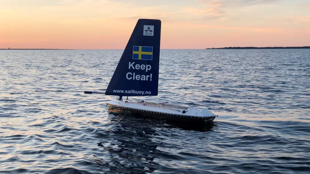 En autonom seglande boj med en blå segelfena som har texten 'Keep Clear!' och en svensk flagga, flyter på en vågig havsyta. Vattenstänk syns i förgrunden, och i bakgrunden syns en kustlinje med byggnader och vindkraftverk under en klar blå himmel.