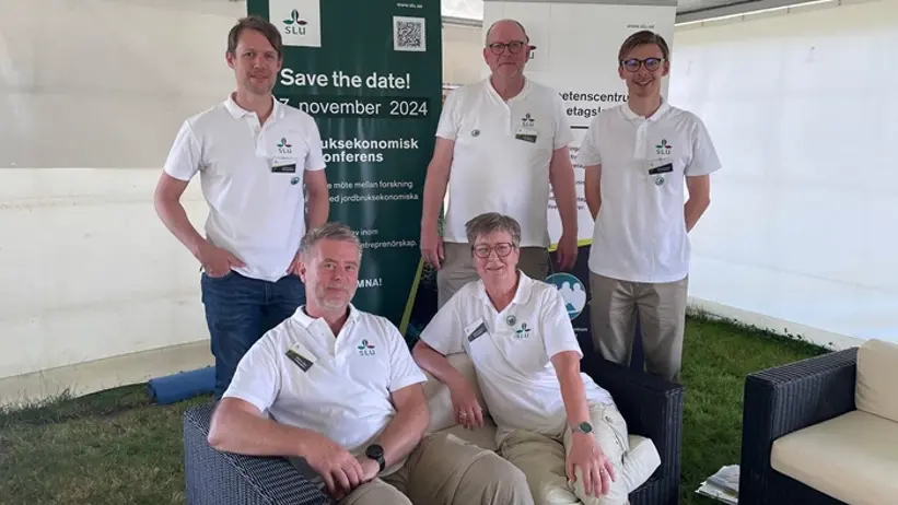 Fem personer står och sitter i en soffgrupp, inne i ett vitt mässtält