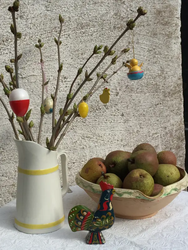 Ett träbord står utomhus, framför en vit husvägg. På bordet står ett påskris, en svart påsktupp i trä och en keramikskål med runda päron.