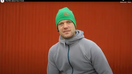 En man i grå sportjacka och grön mössa med orange logotyp sitter framför en röd trävägg och tittar mot kameran. Han bär ett headset i örat, och bilden är hämtad ur en film om SLU:s arbete med livsmedel.