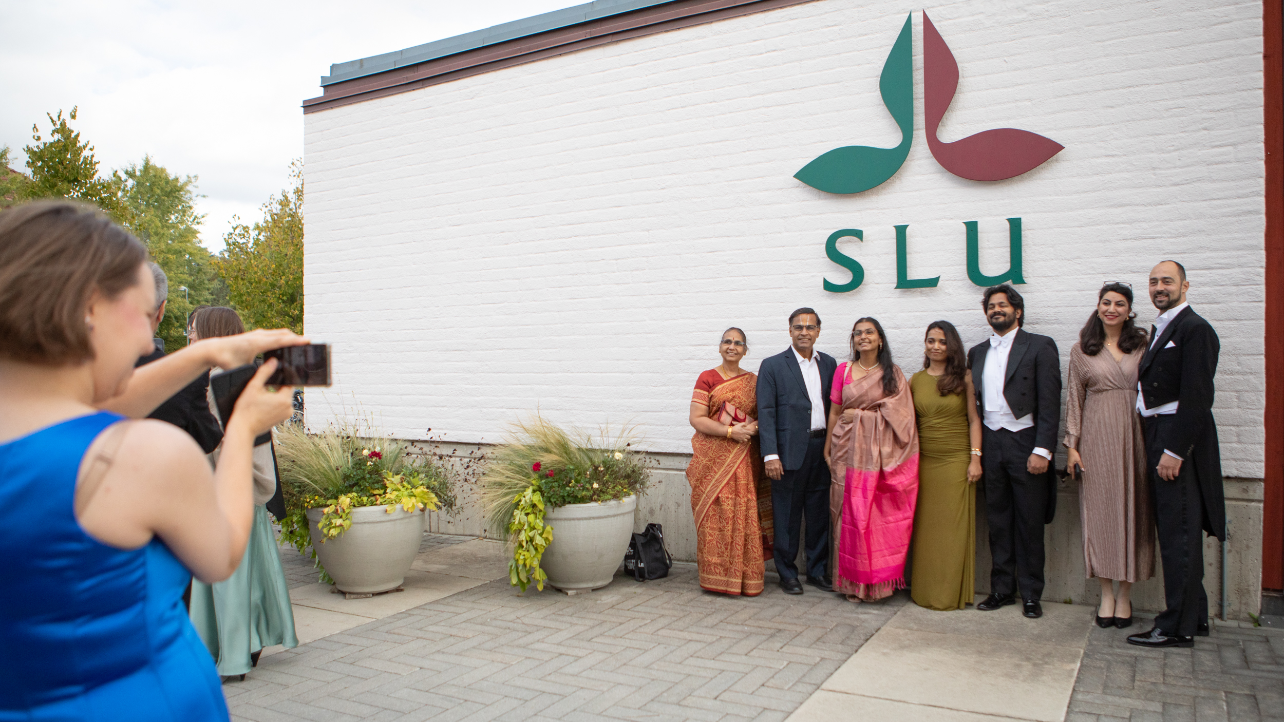 A person takes a photo of a group of festively dressed people standing against a wall with a big logotype on. 