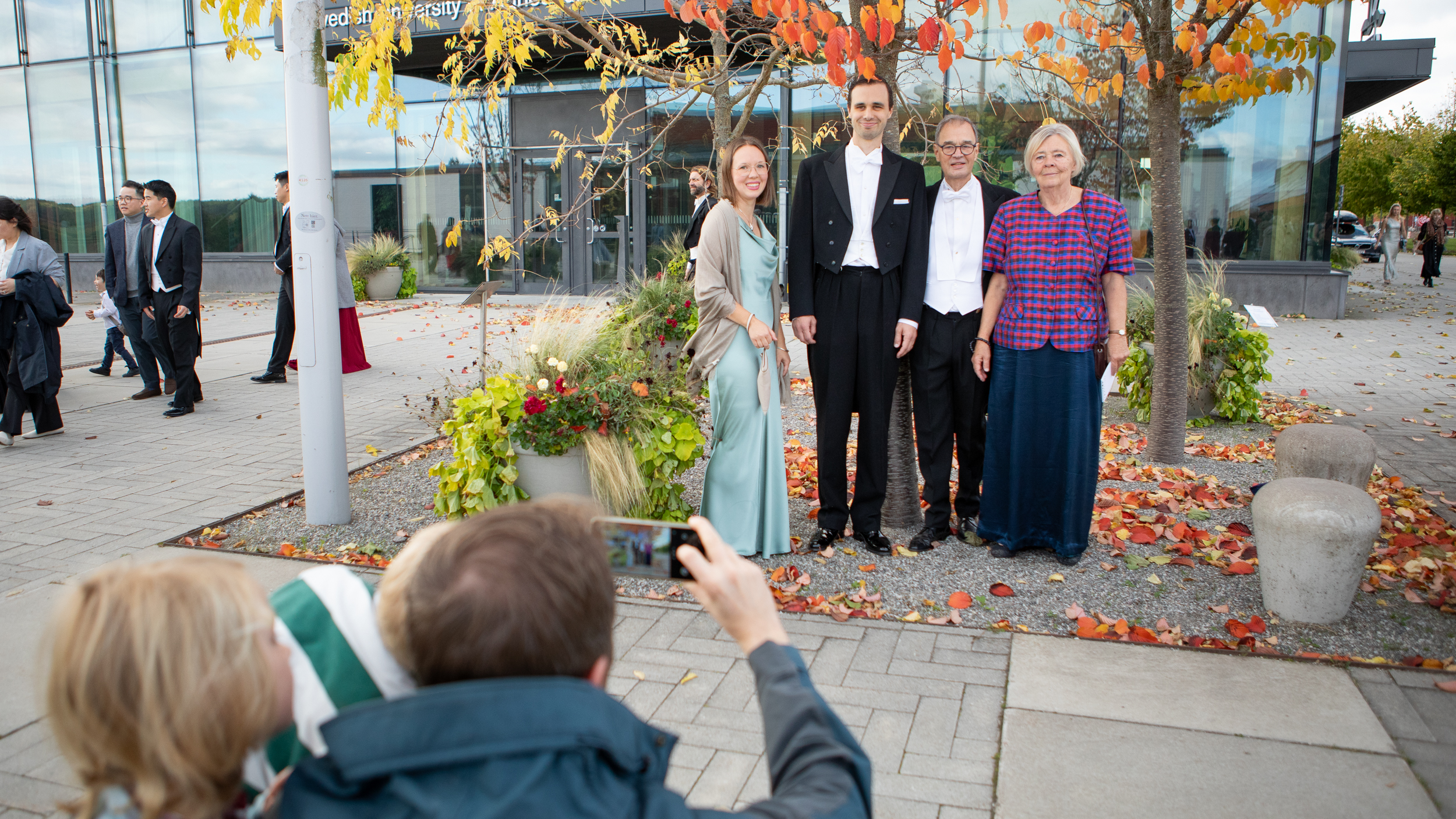 Four festively dressed persons standing below a few trees on a square having their picture taken.