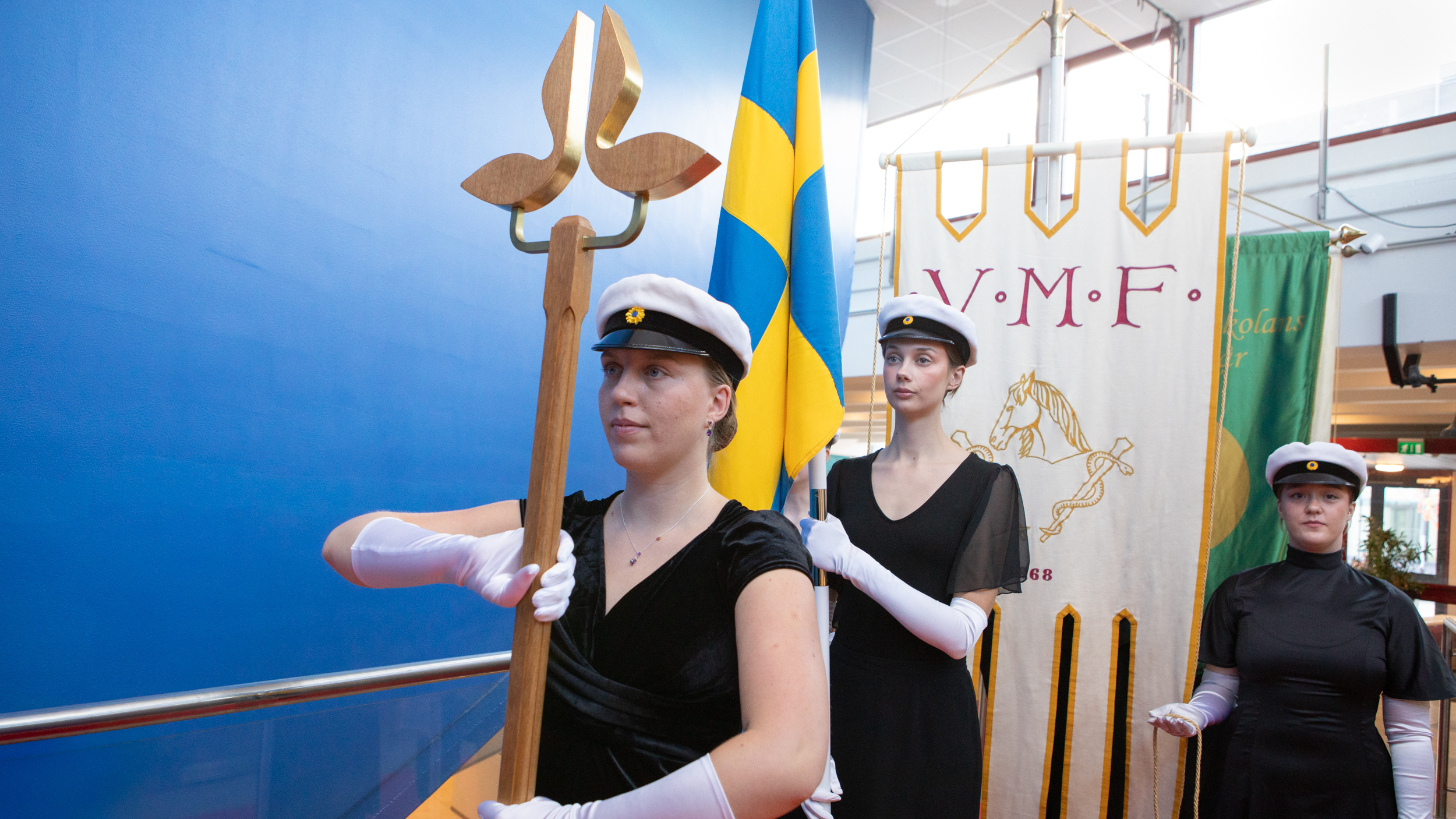 Students dressed in formal attire and wearing graduation caps stand in a row, holding flags. The student at the front carries a wooden staff.