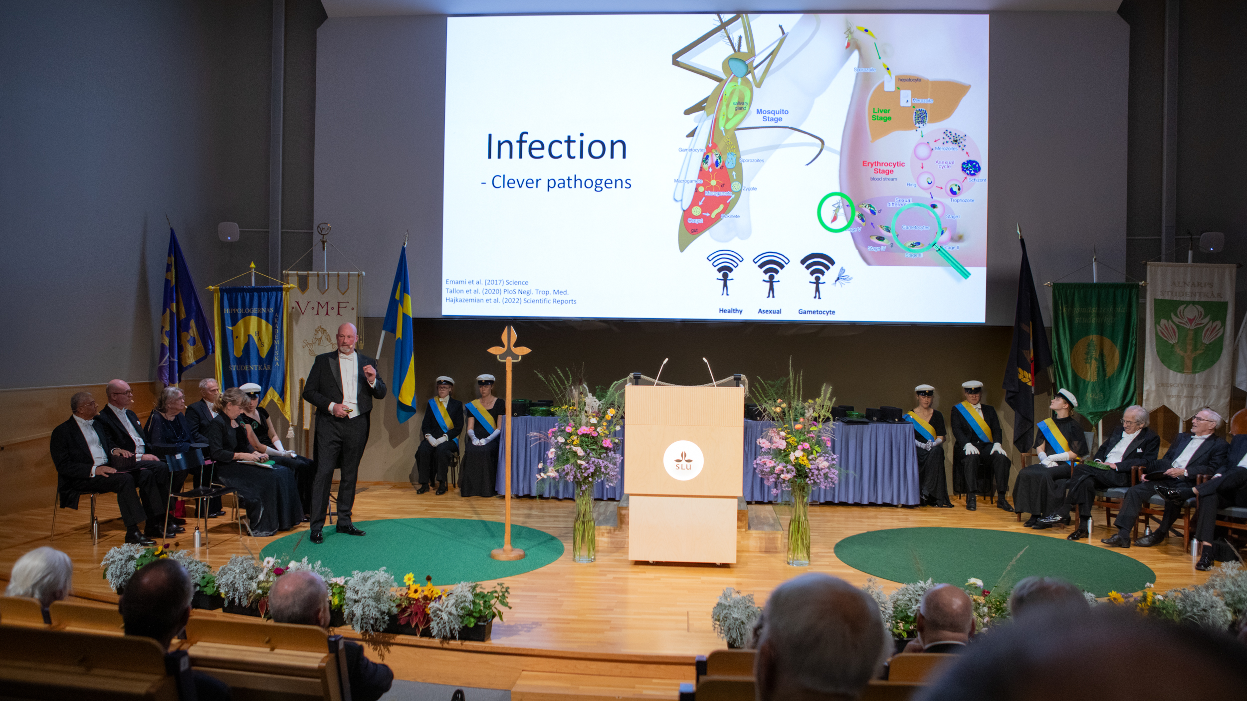 A person in a tailcoat stands on a decorated stage, surrounded by rows of seated people. Projected on the wall behind is a scientific image and the text "Infection – clever pathogens".