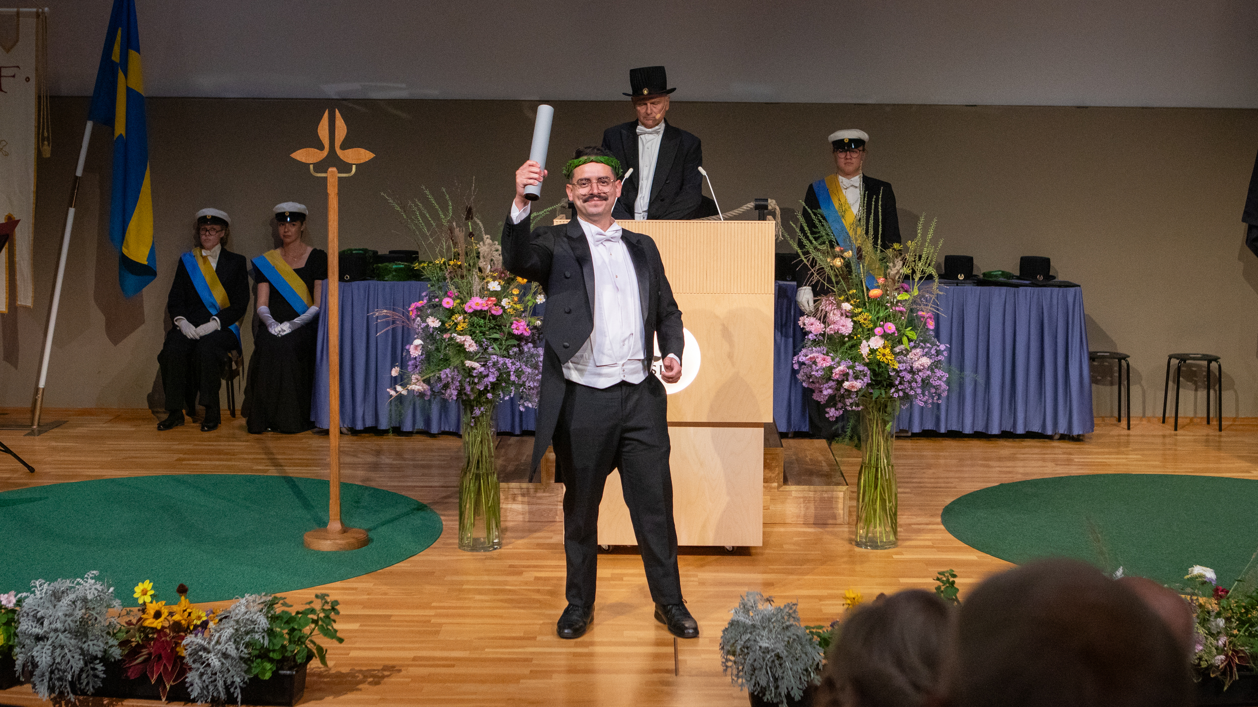A smiling man wearing white tie and a laurel wreath holds up a rolled diploma in a stage.