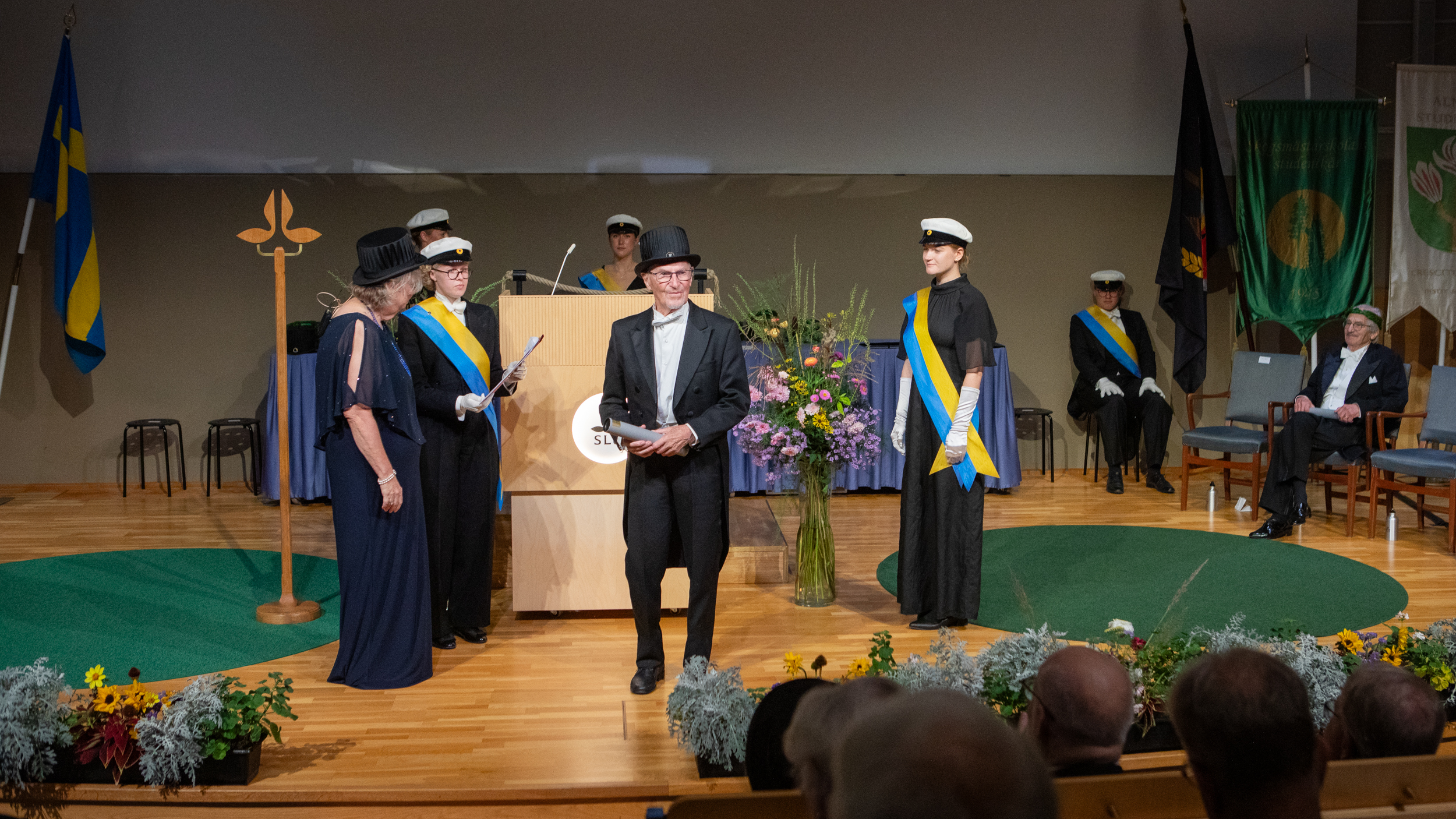An elderly man dressed in white tie and a doctoral hat standing on a stage surrounded by people in student hats and blue-and-yellow silk sashes. 